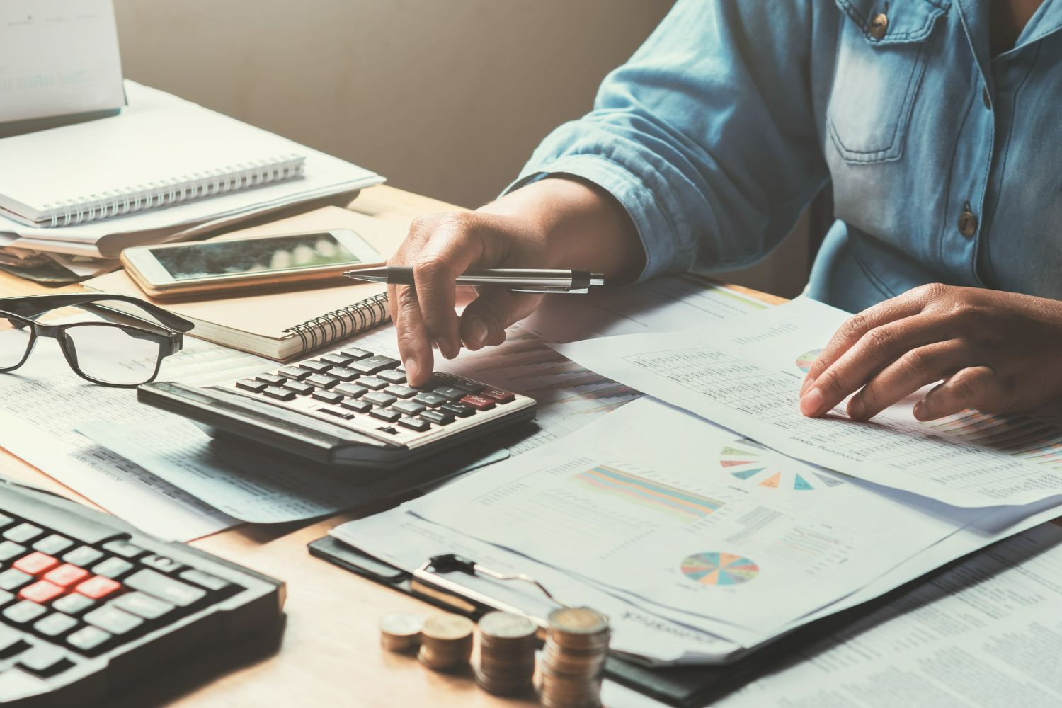 Person calculating with a calculator on a desk with paperwork, coins, and a phone.