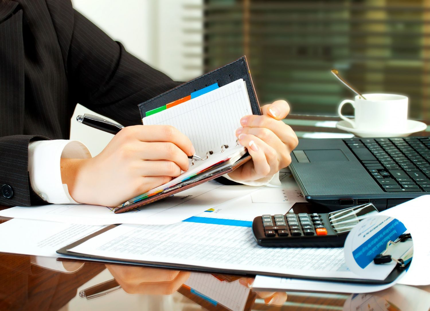 Person in suit writing in planner at desk with laptop, calculator, coffee cup, and documents.