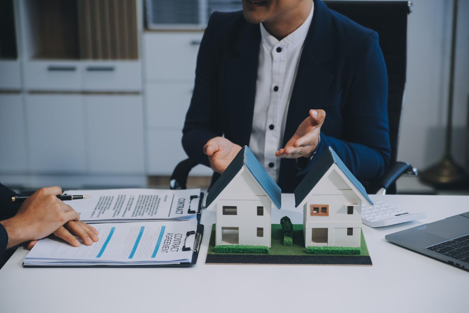 Real estate agent explaining document to a client, model homes and laptop on the table.