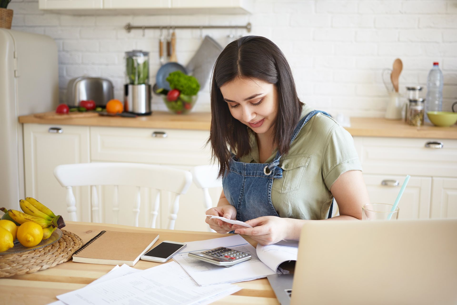 A woman is sitting at a table in a kitchen using a calculator and a laptop.