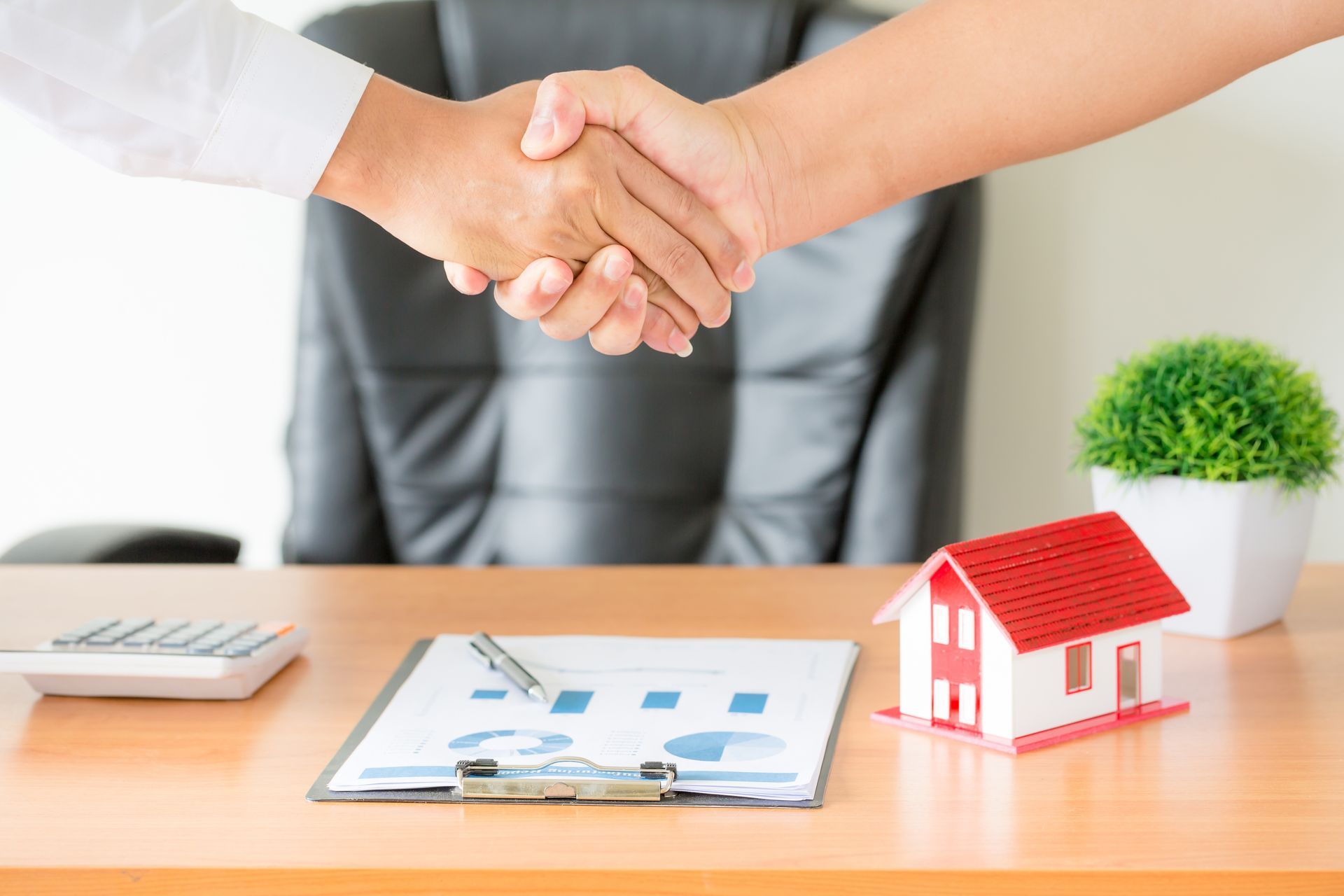 Handshake over desk with house model, tablet, calculator, and potted plant, suggesting a real estate deal