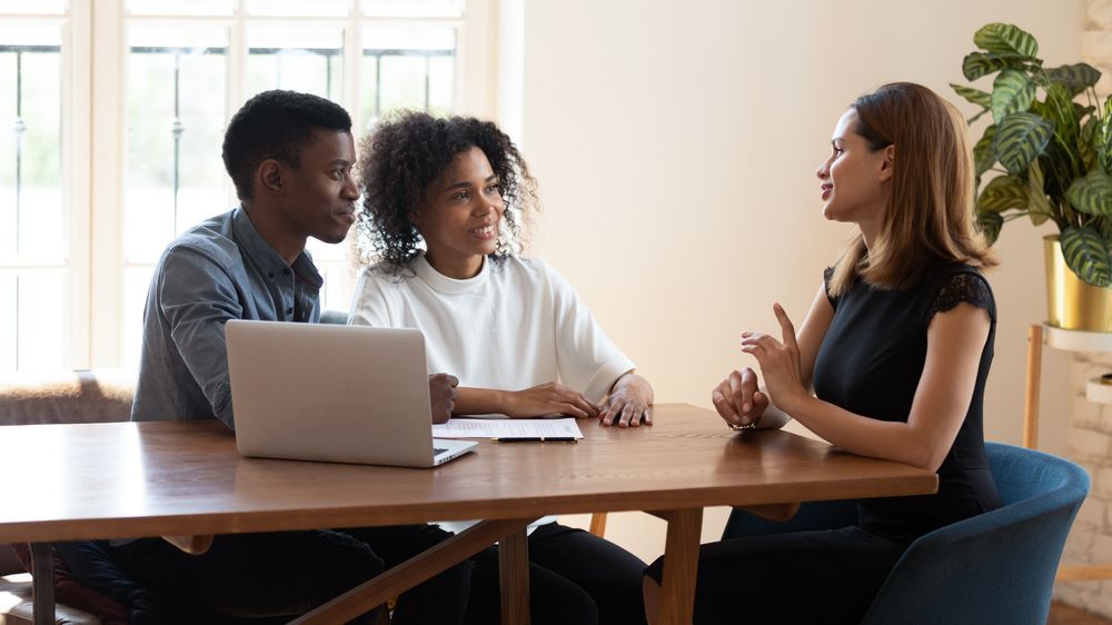 Couple at a table listening to a woman gesturing. Laptop and papers are on the table.