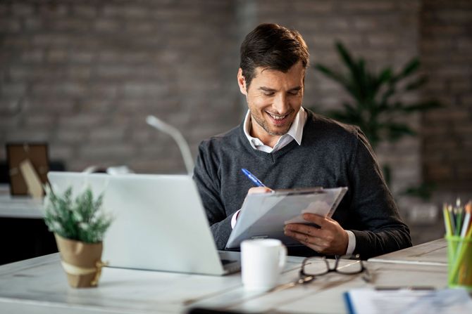 A man is sitting at a desk with a laptop and a clipboard.
