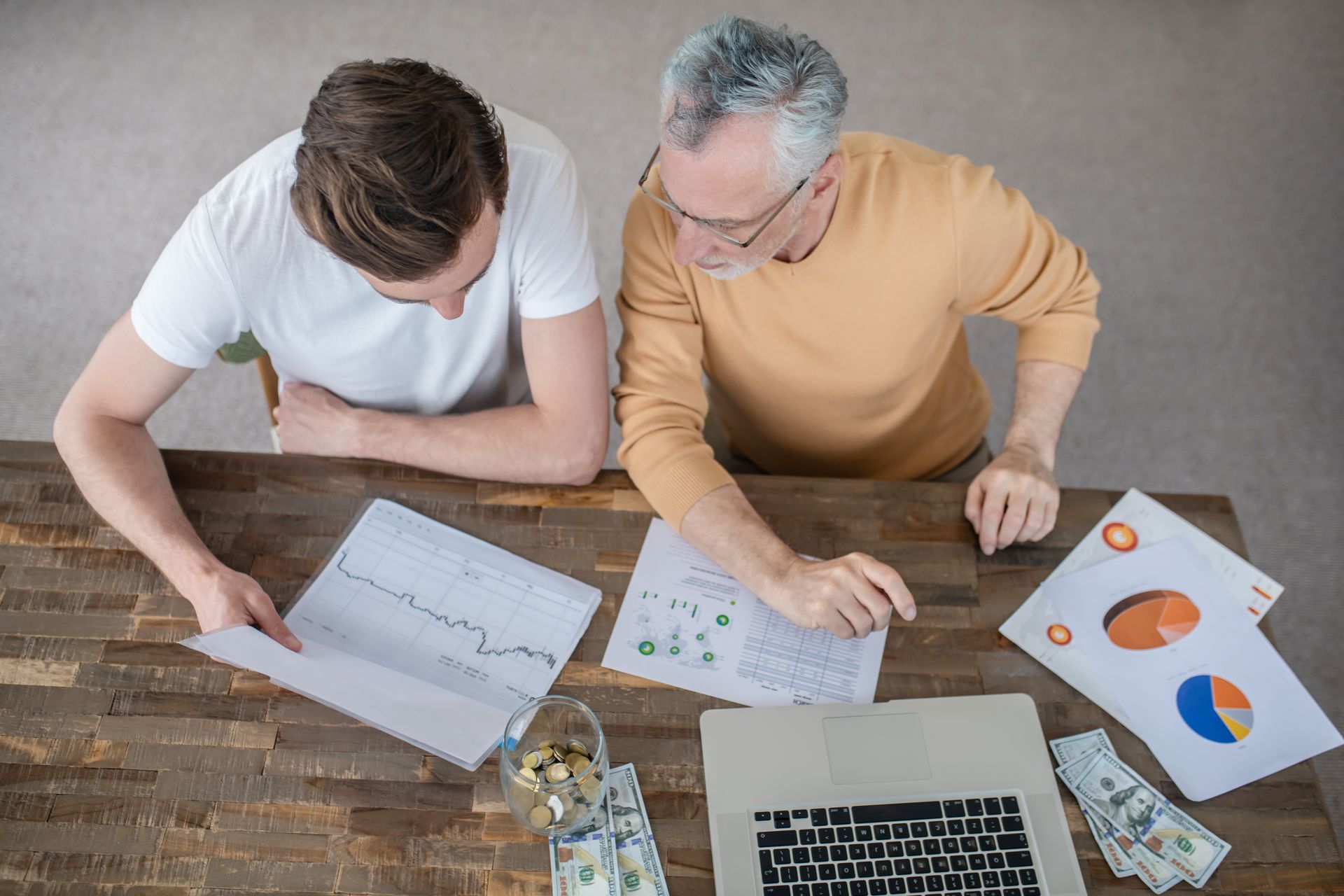 Two men are sitting at a table looking at papers and a laptop.