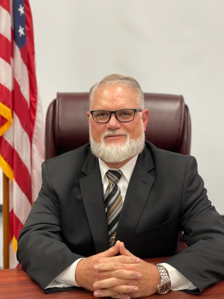 a man in a suit and tie is sitting at a desk with his hands folded in front of an american flag .