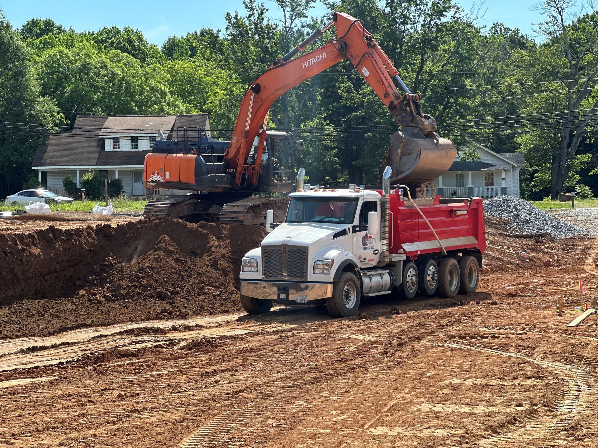 Excavator loading a red dump truck on a construction site; residential neighborhood in background.