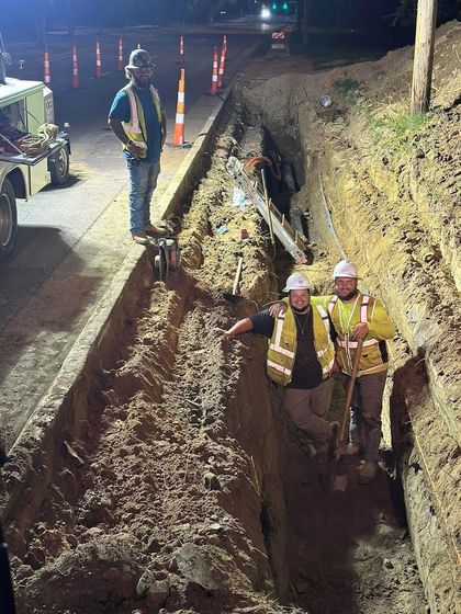 Construction workers in trench on street at night, wearing vests.