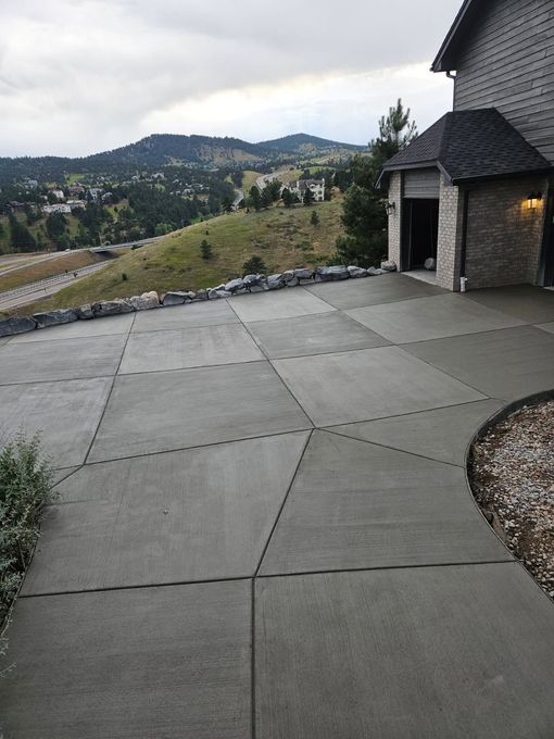 A concrete patio featuring a geometric tile pattern, set against a mountain landscape with a house structure on the right.