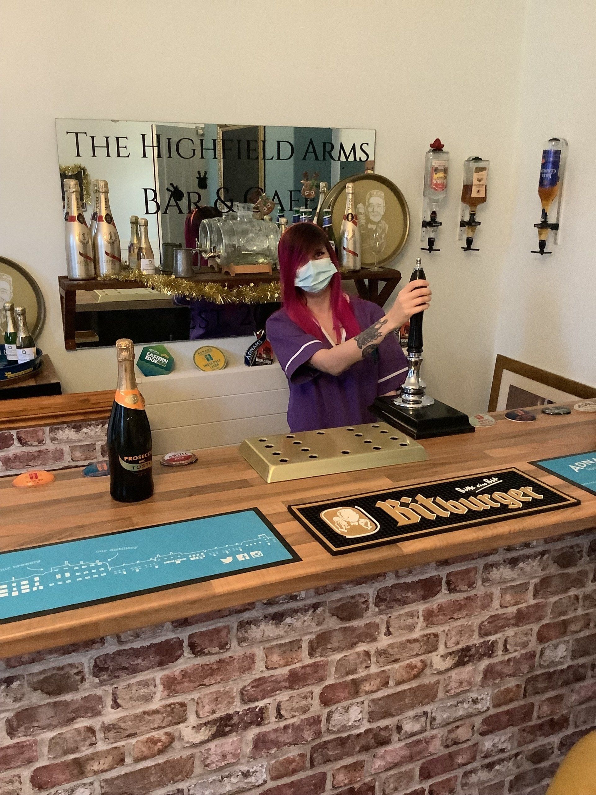 A woman wearing a mask is standing behind a bar
