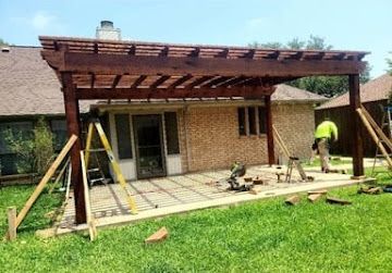 A worker constructs a wooden pergola over a brick house patio on a sunny day.