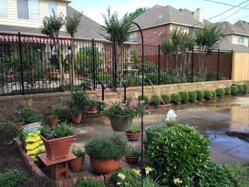 A backyard patio features potted plants, a stone retaining wall, and a black metal fence separating it from a house.