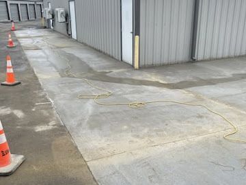 A patch of fresh concrete fills a trench in a gray parking lot next to a metal building, with orange safety cones nearby.