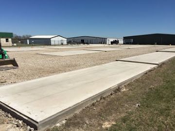 Newly poured concrete pads sit in a gravel yard under a clear blue sky, with commercial buildings in the background.