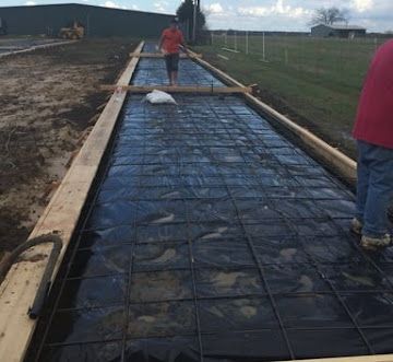 Two people walk on a wooden formwork structure over a black plastic-covered ground with a wire mesh grid for concrete.