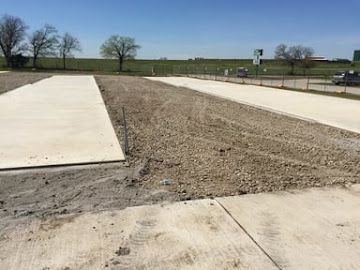 Two concrete slabs are under construction, separated by a gravel area, set against a sunny, open outdoor field.