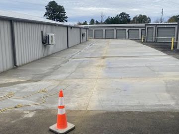An orange traffic cone stands on asphalt in front of a long, gray metal self-storage facility.