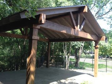 A wooden outdoor pavilion with a gabled roof supported by sturdy posts, located in a park-like setting with green trees.