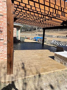 A wooden pergola covers a tan stone patio adjacent to a brick house wall, overlooking a dry, outdoor landscape.