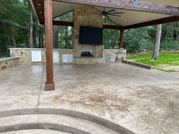 An outdoor stone patio with a roofed fireplace, television, and built-in grill area, viewed from a set of curved steps.