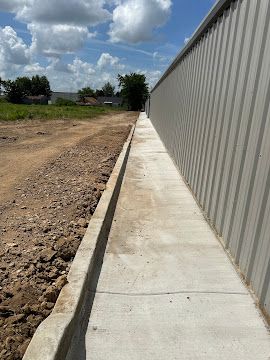 A concrete sidewalk runs along the exterior wall of a metal building next to a dirt construction site under a blue sky.
