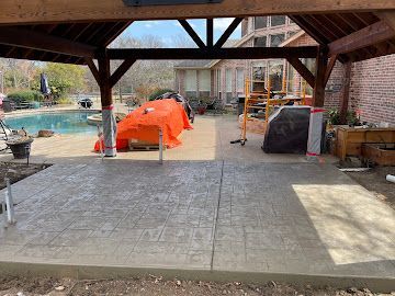 A stamped concrete patio under a wooden pavilion next to a swimming pool and house.