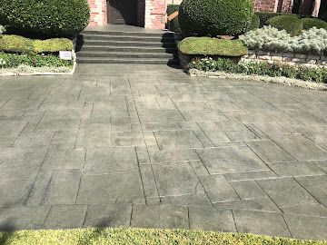 A wide view of a paved entryway leading to stone steps and a front door, surrounded by manicured shrubs.