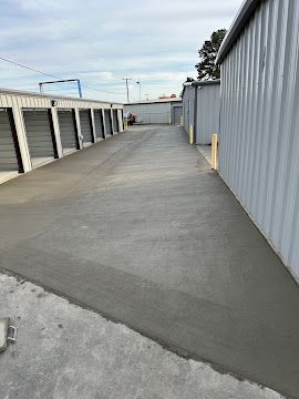 A long, narrow outdoor corridor between rows of grey metal self-storage units with a freshly poured concrete floor.
