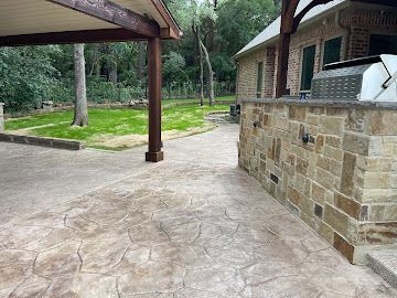 A backyard stone patio with an outdoor kitchen, shaded seating area, and a view of a lush green lawn.