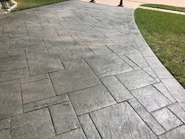 A grey, stamped concrete driveway with a rectangular tile pattern, bordered by green grass.