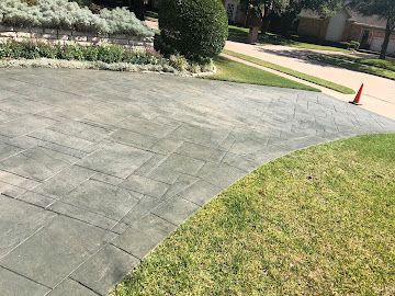 A paved residential driveway made of gray stone tiles, bordered by a green lawn, shrubs, and a small orange traffic cone.