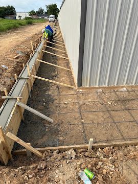 Workers pour concrete into wooden forms alongside a metal-sided building at a construction site.