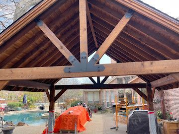 A wooden pavilion frame with prominent black metal hardware at the beam joints, set in a backyard near a swimming pool.