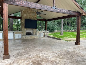 Outdoor covered patio with stamped concrete, a stone fireplace featuring a mounted TV, and a backyard view.