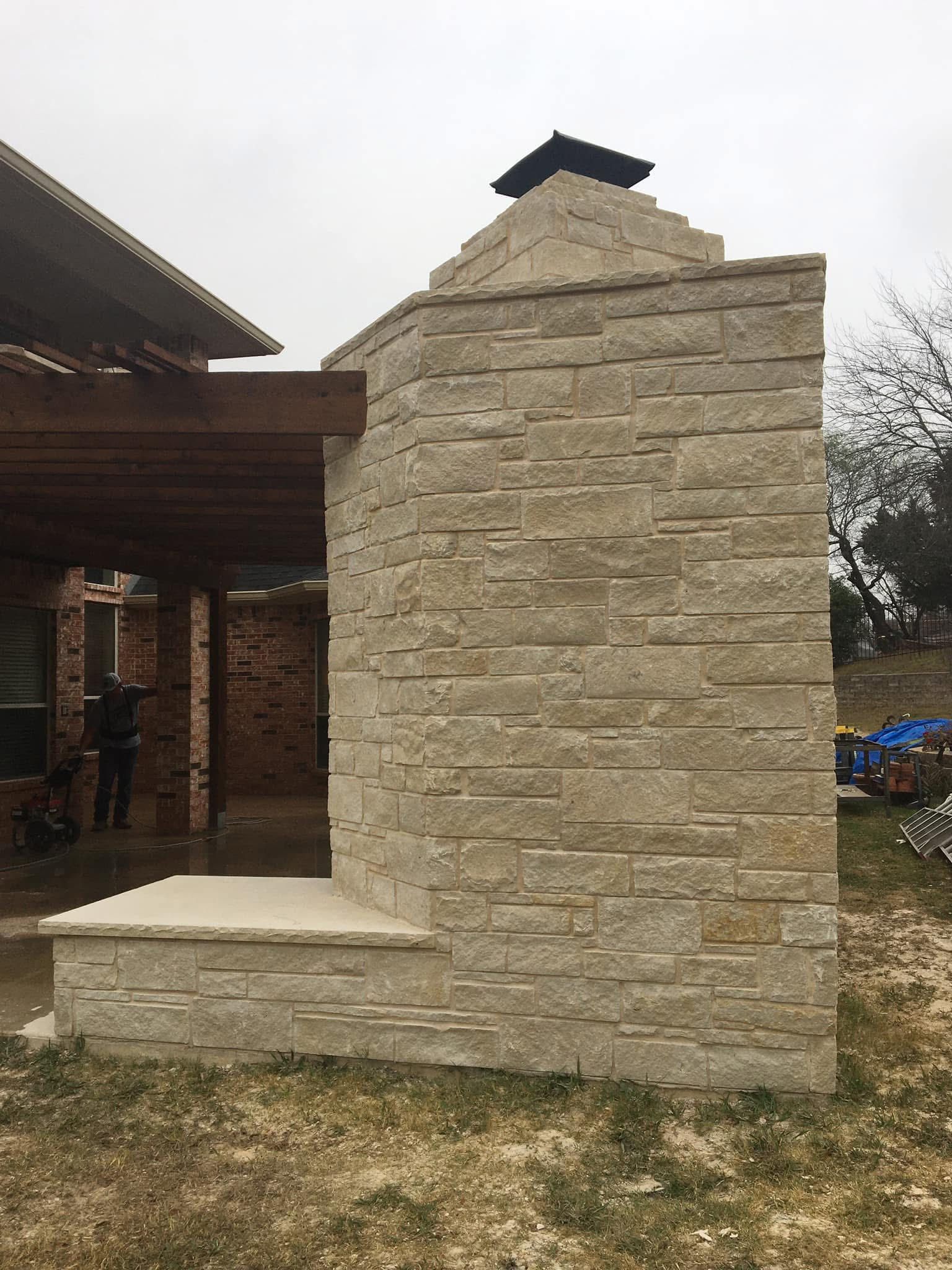 A tan stone outdoor fireplace attached to a wooden patio structure under an overcast sky.