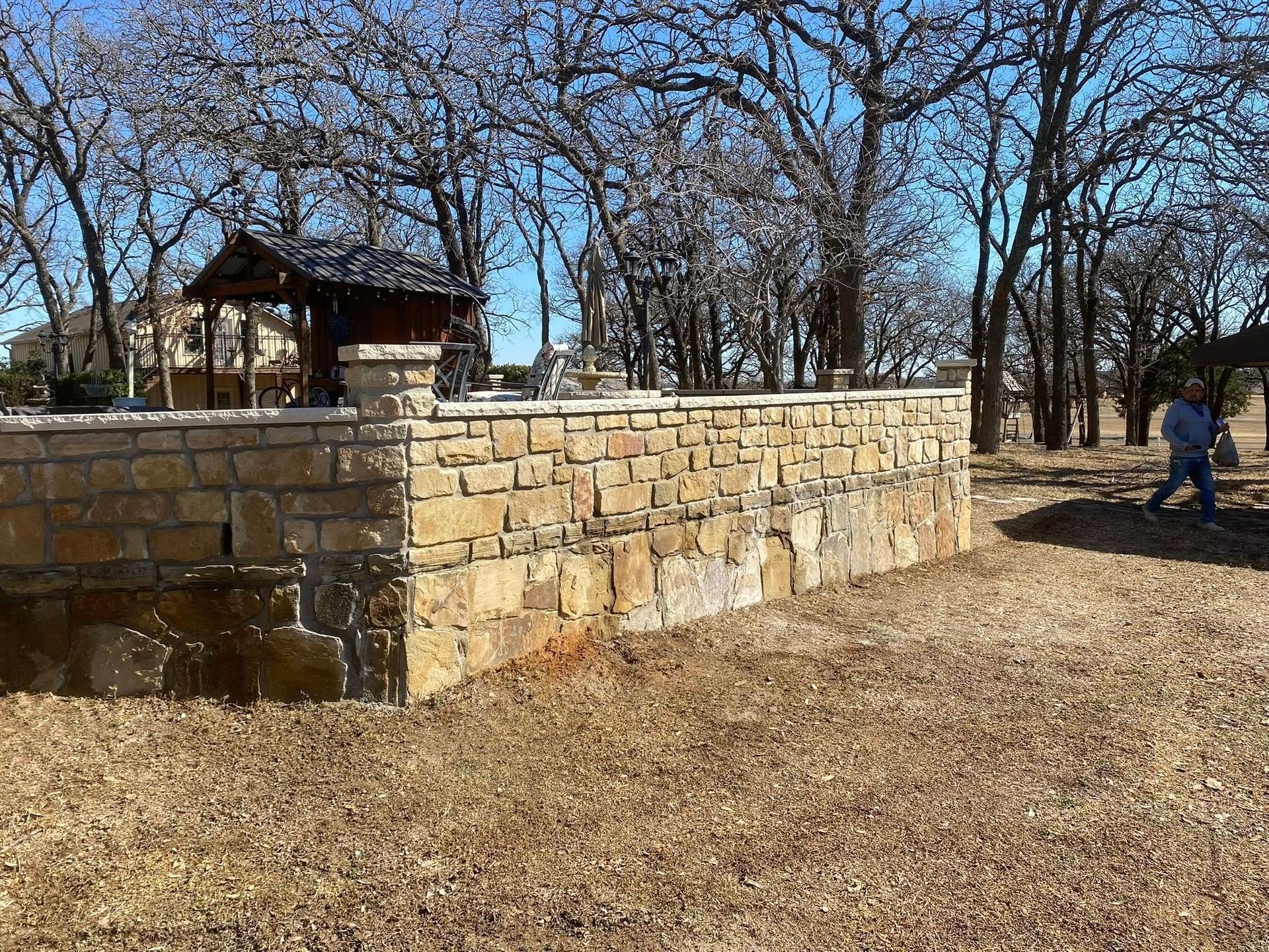 A stone wall surrounds a yard with trees and a small structure, with a person walking in the background.