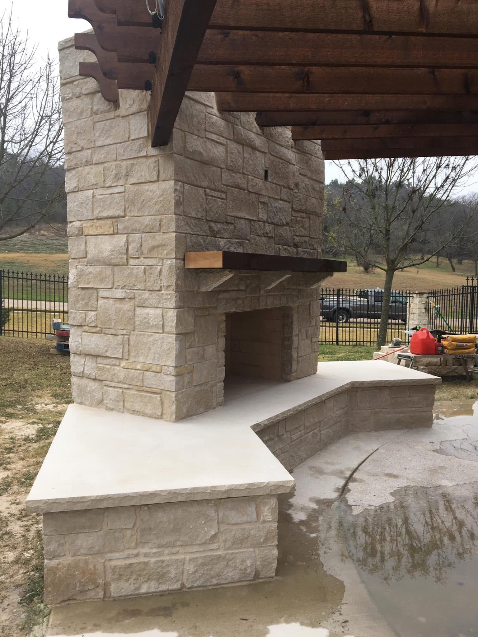 Outdoor stone fireplace with a wooden mantel and bench seating under a pergola in a yard.