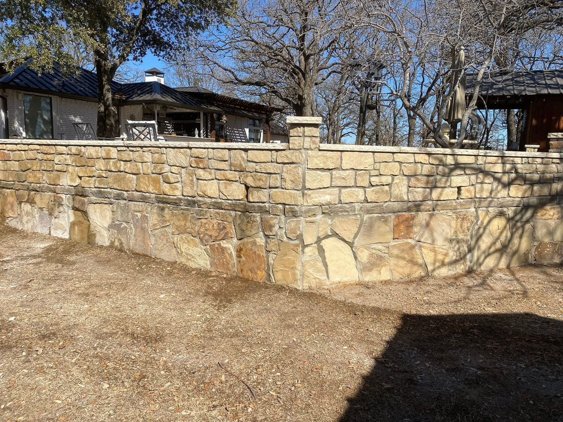 A stone retaining wall built in two styles with a house and bare trees in the background under a clear blue sky.