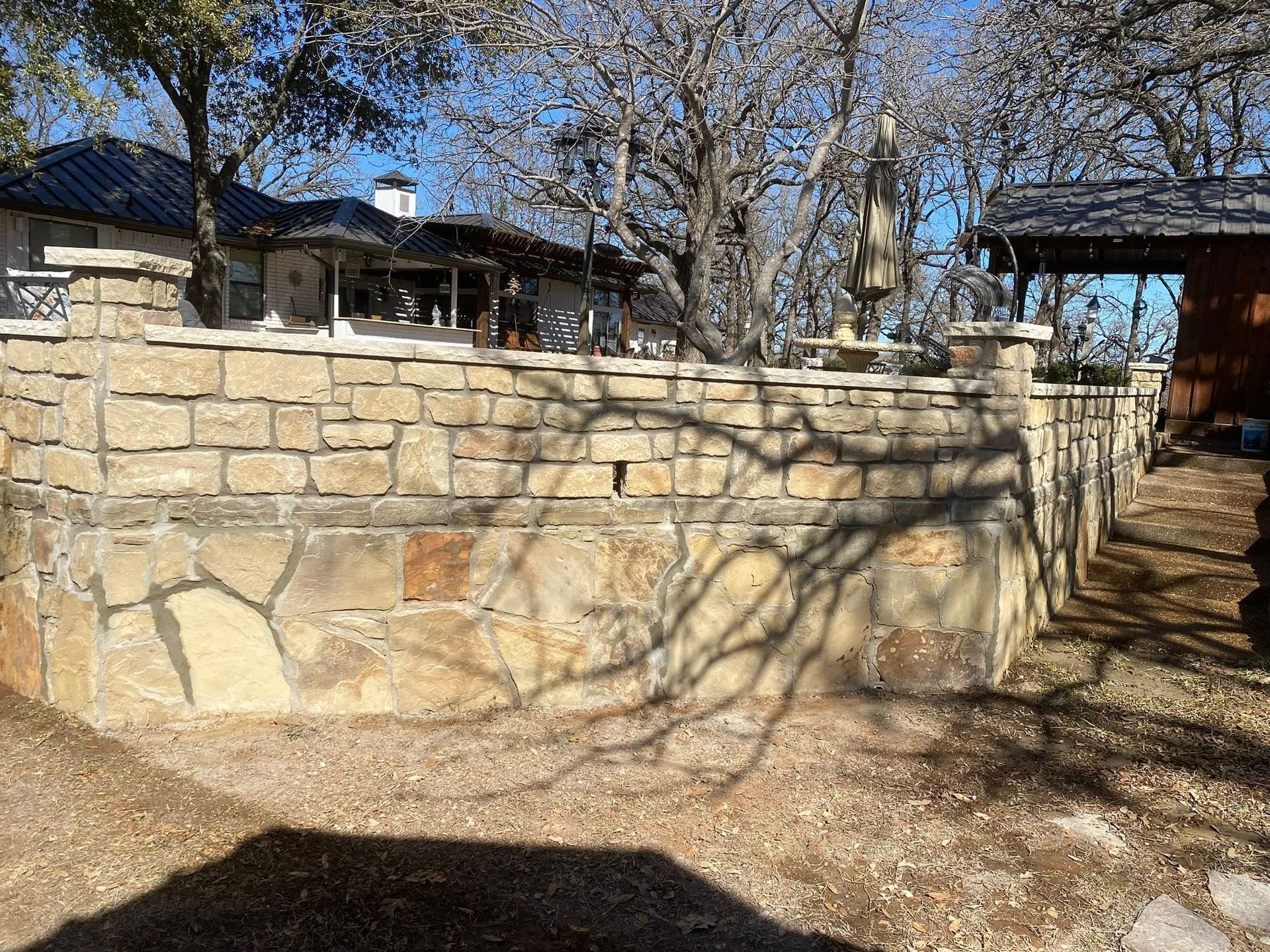 A stone retaining wall in a yard with trees, a house in the background, and a patio structure under a sunny sky.