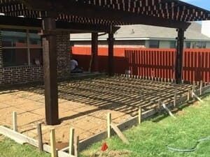 A construction site for a backyard patio featuring wooden forms, a prepared sand base, and an overhead dark wood pergola.