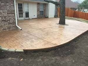 A tan, stamped concrete patio with a curved edge surrounds a tree next to a brick house with a glass door.