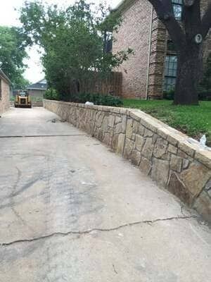A concrete driveway runs alongside a tan stone retaining wall beside a brick house and tree.