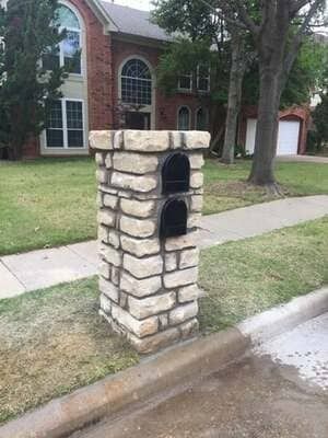 A stone mailbox pillar with two black mailboxes stacked vertically, situated on a lawn in front of a brick house.