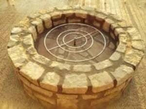 A round stone fire pit with a concentric metal burner ring installed inside on a gravel surface.
