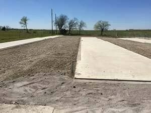 Rows of newly poured concrete pads sit separated by strips of gravel in an open, grassy field under a clear blue sky.