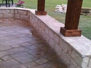 A stone patio with a curved retaining wall and wooden support posts for a pergola, with a green lawn in the background.