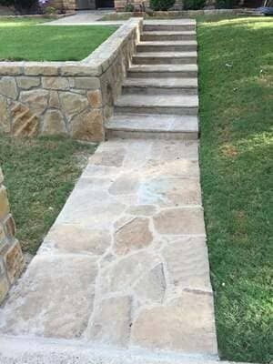 A stone walkway leads to a set of stone steps surrounded by green grass and a stone retaining wall.