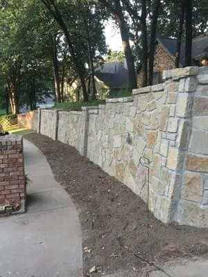A curved stone retaining wall lines a concrete walkway beside a patch of dirt, with trees and houses in the background.