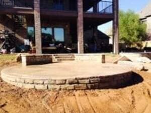 A circular concrete patio with a stone retaining wall edge, situated in a backyard under construction near a house.