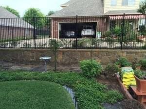 A suburban backyard with a stone retaining wall, a metal fence, a birdbath, and a house with an open garage in the distance.