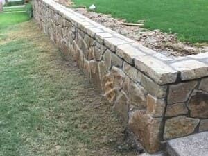 A low, tan stone retaining wall borders a grassy lawn, viewed from the side at ground level.